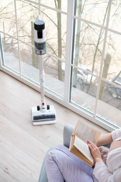 Close-up of vacuum cleaner tube. Female figure in chair by panoramic window with open book. Rechargeable vacuum cleaner with dust collector on handle. Vertical shot.