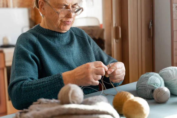 一位视力不好的老年男子坐在厨房里织毛衣 桌上有蓝色 棕色和灰色的线组成的球 专注于脸部 — 图库照片