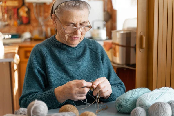 一位白发苍苍 头戴发带的老妇人坐在家里用针织针头织毛衣 桌子上有毛线 背景上厨房的水泡图像 — 图库照片