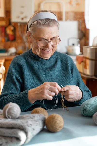 一位快乐的八十岁老妇人坐在桌旁 用毛线编织针头 大眼镜的视觉 老年人的业余爱好 — 图库照片