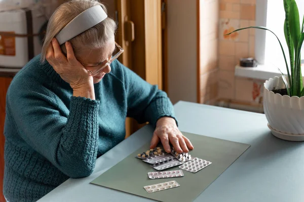 Üzgün yaşlı bir kadın bir odada oturuyor ve büyük miktarda ilaca bakıyor. Yaşlı bir kadının birçok hastalığı vardır ve tedaviye ihtiyacı vardır. Yaşlılıkta sağlık kavramı.
