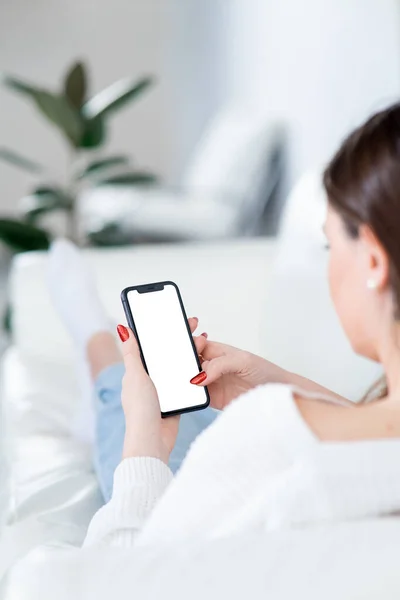 Modern akıllı telefonlu genç bir kadın kanepede yatıyor. Işığın arka planı ve sıcak iç mekan. Reklamınız için telefon ekranı modeli.