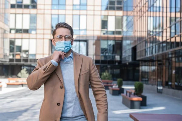 Serious business man in glasses and mask on street, against background of modern business center, covid-19 pandemic, concept of protected business.