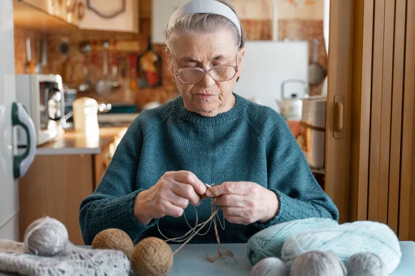 An elderly lady of 80 years old is engaged in an interesting hobby, knitting a brown jacket with knitting needles. Hobbies that you can do at home. A blurry image of the kitchen on the background.