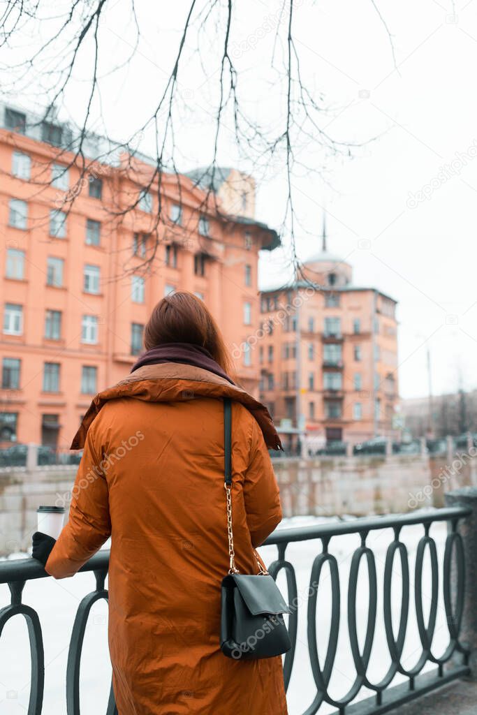 Enjoying the view of the river and the city, the woman stands with her back. Relaxation during the walk. Warm clothes in the cold season. An iron barrier to safety. Vertical photo.