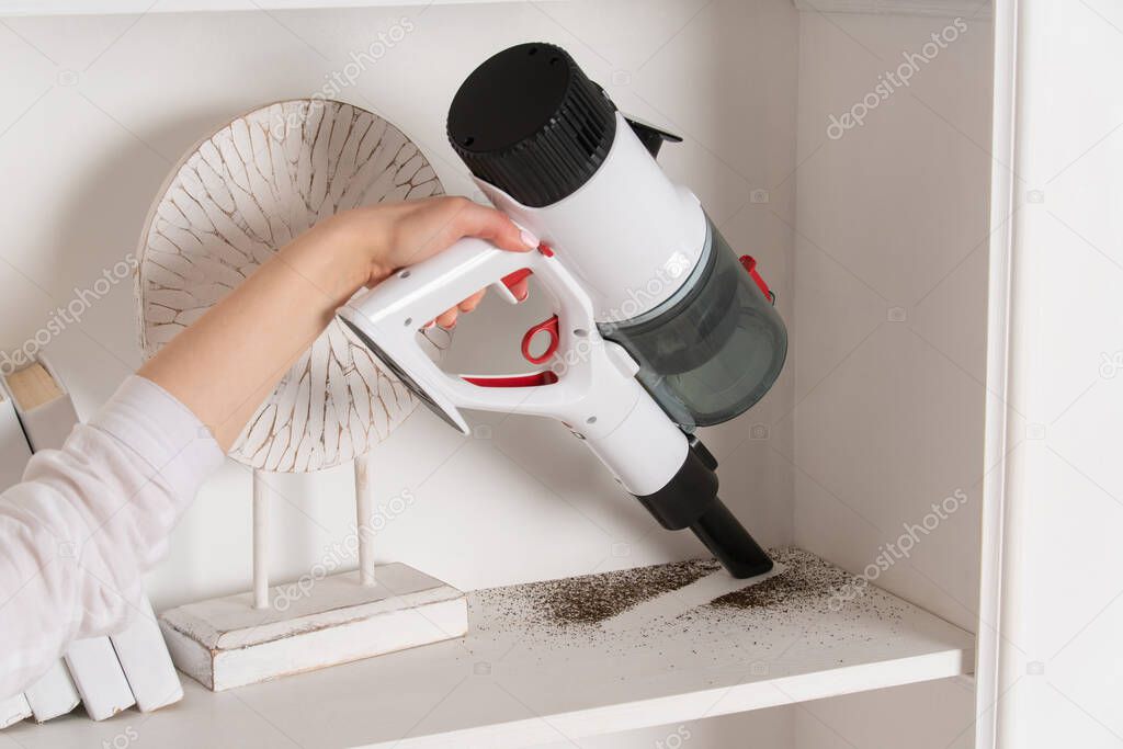 Woman vacuums shelves in room. Women's hand with portable vacuum cleaner. Rolled up sleeves. Items on shelf. Cleaning. Close-up of Dust collector.