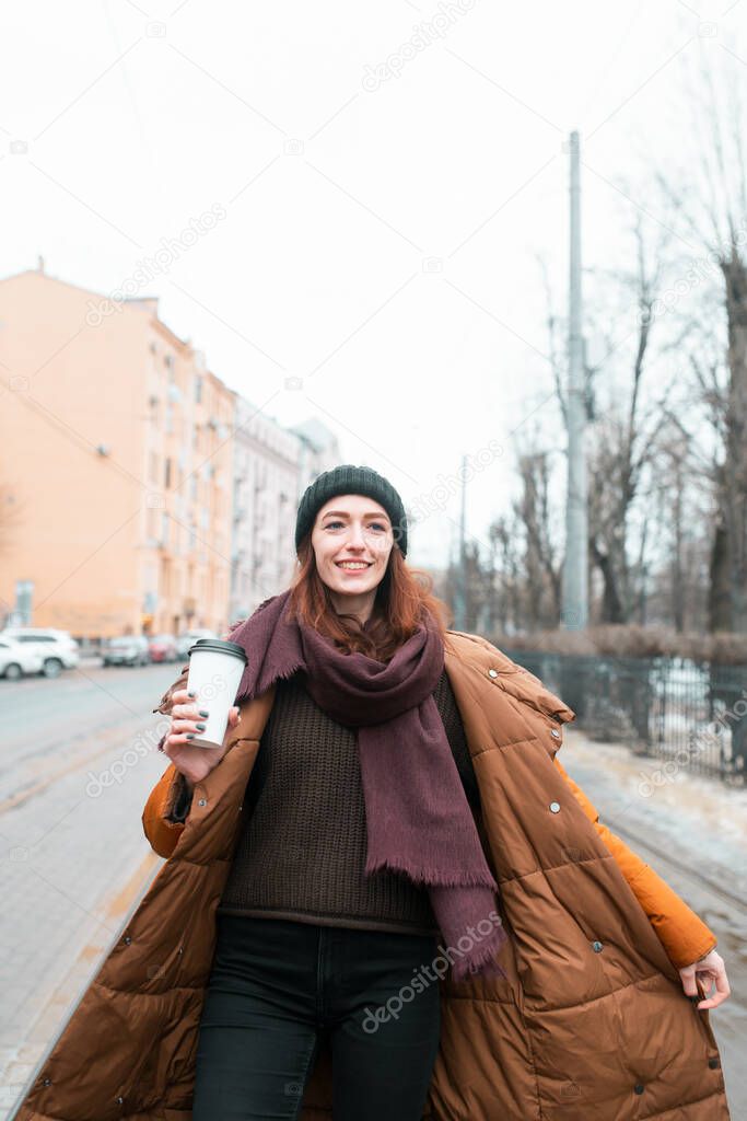 The redhead smiles and walks down the street with a happy expression on her face. In an orange long jacket and a mug of coffee. Looks away. Blurry image of the city.