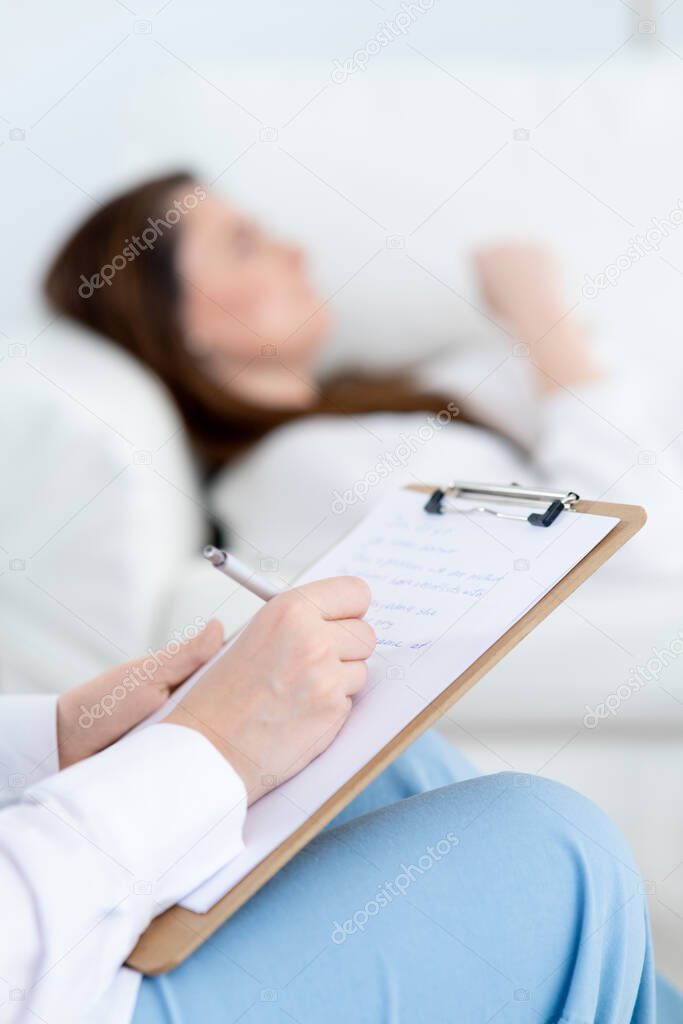 Close-up of the therapist hands writing in the clipboard. The patient lies on and talks about his problems. Professional help of a psychotherapist. Bright office. Mental health.