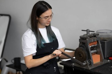 Siyah iş önlüğü, beyaz tişört ve güvenlik gözlüğü takan mutlu genç kadın. Yuvarlak gümüş. Orijinal Mücevher Atölyesi.