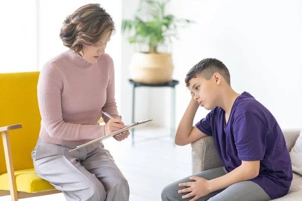 Upset teenager holding his head. Woman psychologist writes on clipboard. Appointment with teenage psychologist. School psychologist.