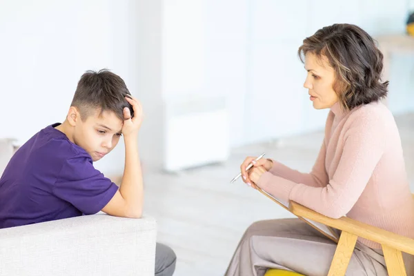 Upset teenager holding his head. Middle-aged woman psychologist talks to him. Mental health. School psychologist.