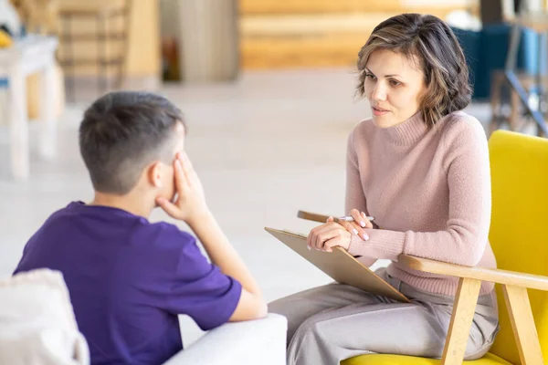 Upset teenager holding his head. Middle-aged woman psychologist talks to him. Mental health. School psychologist. Against background of light office.