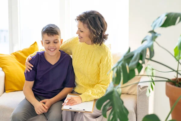 Woman, school psychologist, hugs and supports teenager while sitting on sofa by window. Cozy and light interior of office. Monstera plant in blurred foreground.