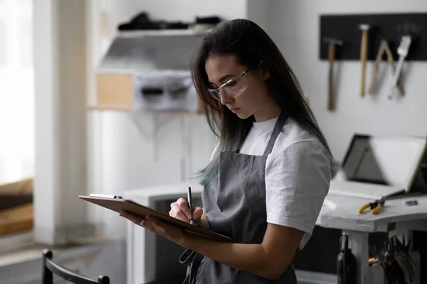 Young woman jeweler in gray apron holds clipboard in her hands. Makes sketches for new products. Against background of workbench with tools in jewelry workshop.