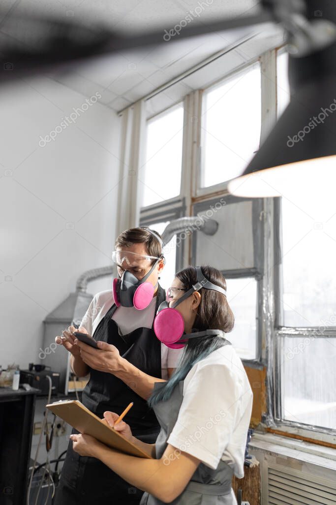 Young couple of jewelers in black aprons, white T-shirts and pink respirators planning day. small workshop. Making handmade jewelry. Blurred foreground.