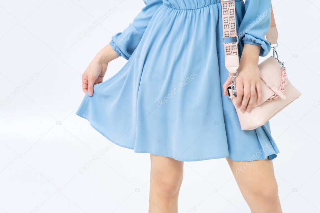 Close-up of the bottom of a light blue summer dress on a young woman, slender bare legs. A light pink handbag with a chain. White background.