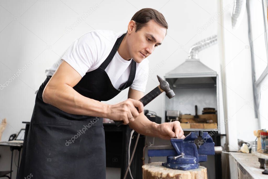 Young man in black apron holds hammer and metal blank. Straightens the workpiece. Hits with hammer. Workshop interior in the background