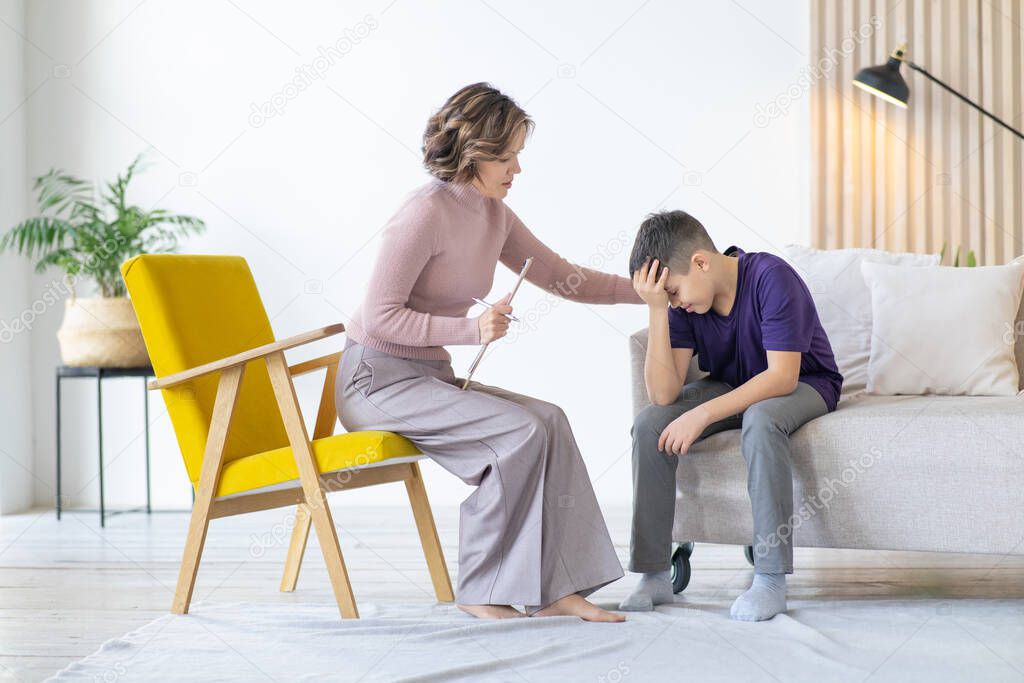 Counseling adolescent psychologist calms and holds adolescent by shoulder. Boy in purple t-shirt is worried and holds his head with his hand, while sitting on couch in minimalistic room. Mental health.