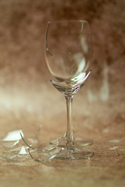 İki kırık şarap kadehi solgun bir kum zeminine karşı biri diğerinin önünde, altında kırık cam parçaları olan.