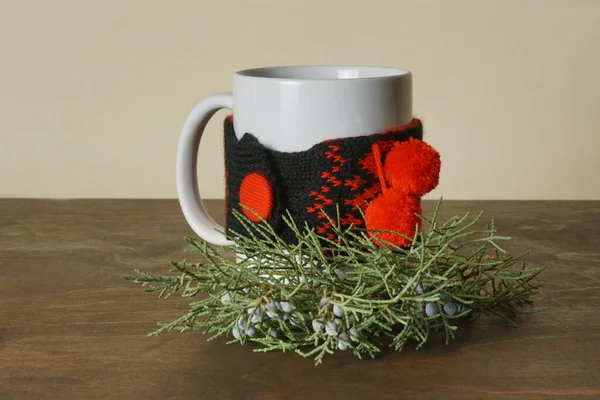 Beyaz seramik bir kupa, kahverengi bir masanın üzerine örülmüş bir kolla. Arka planda yeşil bir ardıç dalı, mavi-yeşil meyveli..