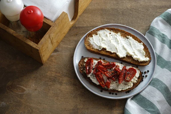Yuvarlak ev yapımı ekmek, kurutulmuş domates ve baharatlı krem peynirden yapılmış sandviç. Tahta bir masada, beyaz bir tabakta sandviç..