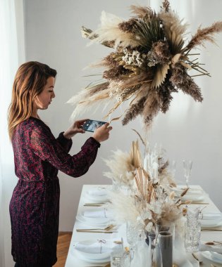 Evde süslenmiş Noel Yemeği Masası 'nın fotoğrafını çeken genç bir kadın. Yemek masasındaki zarif kadın öğle yemeği partisinden önce cep telefonuyla yemek masasına kurulmuş..