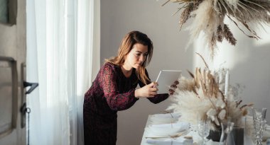 Güzel bir kadın evde süslenmiş bir Noel yemeği masası fotoğrafı çekiyor. Ot ayıklamadan önce dijital tabletle hazırlanmış elbise fotoğraf masasındaki kadın veya doğum günü partisi.