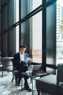 Yakışıklı Asyalı İş Adamı Kafe 'de Belgeler Okuyor. Mavi takım elbiseli ciddi bir iş adamı büyük pencerelerin yanındaki kafede otururken iş raporlarını tutuyor ve analiz ediyor..