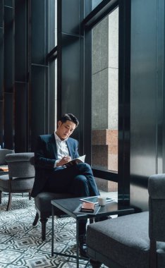 Yakışıklı Asyalı İş Adamı Kafe 'de Belgeler Okuyor. Mavi takım elbiseli ciddi bir iş adamı büyük pencerelerin yanındaki kafede otururken iş raporlarını tutuyor ve analiz ediyor..