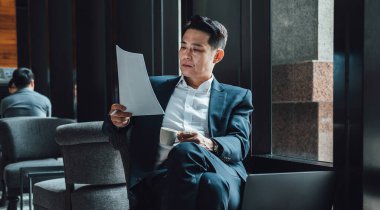 Yakışıklı Asyalı İş Adamı Kafe 'de Belgeler Okuyor. Mavi takım elbiseli ciddi bir iş adamı büyük pencerelerin yanındaki kafede otururken iş raporlarını tutuyor ve analiz ediyor..