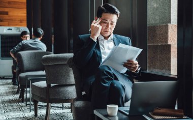 Yakışıklı Asyalı İş Adamı Kafe 'de Belgeler Okuyor. Mavi takım elbiseli ciddi bir iş adamı büyük pencerelerin yanındaki kafede otururken iş raporlarını tutuyor ve analiz ediyor..