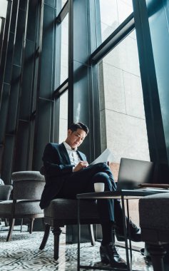 Yakışıklı Asyalı İş Adamı Kafe 'de Belgeler Okuyor. Mavi takım elbiseli ciddi bir iş adamı büyük pencerelerin yanındaki kafede otururken iş raporlarını tutuyor ve analiz ediyor..