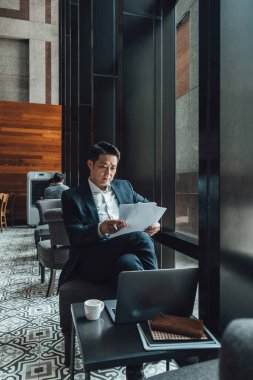 Yakışıklı Asyalı İş Adamı Kafe 'de Belgeler Okuyor. Mavi takım elbiseli ciddi bir iş adamı büyük pencerelerin yanındaki kafede otururken iş raporlarını tutuyor ve analiz ediyor..