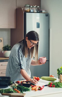Mutfakta çay içerken Sağlıklı Tarifler Arayan Güzel Kadın. Ciddi kadın şef bir deftere yemek tarifi yazıyor ve öğle yemeği için taze sebzelerle dolu bir masada dururken elinde bir fincan kahve tutuyor.. 