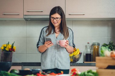 Mutfakta bir fincan çay içerken cep telefonu kullanan mutlu kadın. Gülümseyen kadın şef elinde bir fincan kahve tutarken ve telefonuna mesaj yazarken ya da evde internetten yemek tarifi okurken.