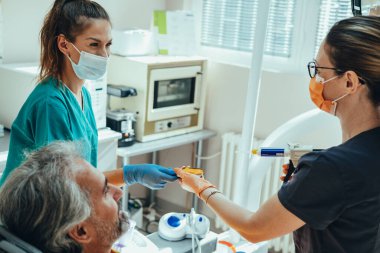 Diş implantı için diş izi almak ya da diş hekiminin muayenehanesinde diş hekiminden oluşan mutlu bir takımla taç yaptırmak. Olgun erkek hasta dişçi koltuğunda otururken dişçi ve dişçi asistanı cerrahi maske ve eldiven takıp diş izi alıyor..