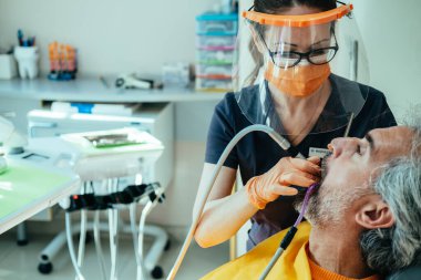 Coronavirus salgını sırasında dişçi muayenehanesinde dişçi muayenesi yaptıran ciddi bir olgun adam. Koruyucu iş kıyafeti giyen kadın diş sağlığı kliniğinde erkek hasta dişlerindeki diş dolgusunu tedavi etmek için diş matkabı kullanıyor..