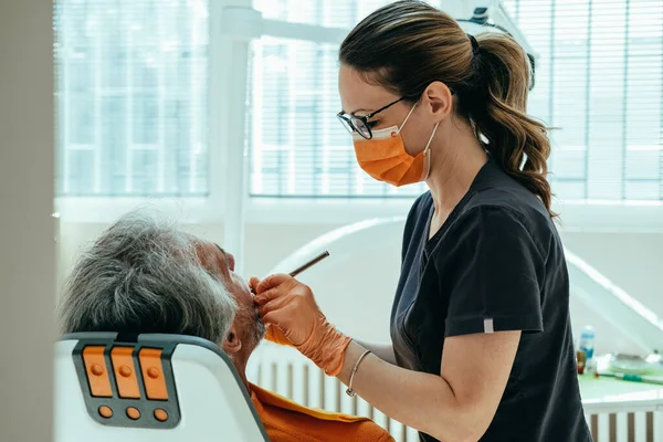 Diş Kliniği 'nde çalışan kadın dişçi. Dişçi koltuğunda tanınmayan bir erkek hasta var. Turuncu koruyucu maskeli ve eldivenli kadın dişçi muayenehanesinde yetişkin hastasını muayene ediyor..