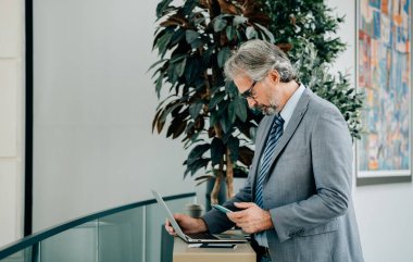 Havaalanında cep telefonu ve dizüstü bilgisayar kullanan Olgun İşadamı Uçağı Bekliyor. Ciddi bir iş adamı dizüstü bilgisayarı, pasaportu, bileti ve bir fincan kahveyle havaalanı terminalinde akıllı telefonunu tutuyor..