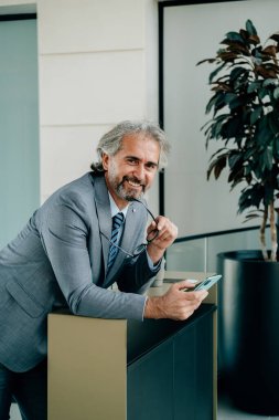 İş yerinde cep telefonu kullanan gülümseyen bir iş adamının portresi. Modern ofis binasındaki bir masaya yaslanıp kameraya bakarken şık gri takım elbiseli yakışıklı ve yakışıklı bir iş adamı akıllı telefonunu ve gözlüklerini tutuyor..