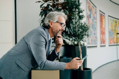 Gözlüklü gülümseyen iş adamı iş yerinde cep telefonu kullanıyor. Zarif gri takım elbiseli yakışıklı bir iş adamı elinde akıllı telefonuyla modern ofis binasında bir masaya yaslanıp bir şeyler düşünürken..