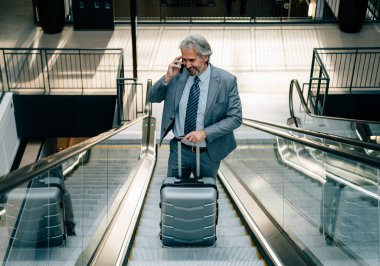 Gülümseyen Olgun İşadamı yürüyen merdivende duruyor ve cep telefonuyla konuşuyor. Havalimanında bavuluyla yürüyen merdivenden çıkarken zarif gri takımlı mutlu iş adamı telefonda konuşuyor.. 