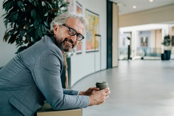 Kahve molası: Gülümseyen bir iş adamının İş yerinde bir fincan kahve içen portresi. Modern ofis binasındaki bir masaya yaslanıp kameraya bakarken zarif gri takım elbiseli yakışıklı, olgun bir iş adamı elinde bir fincan çay tutuyordu..