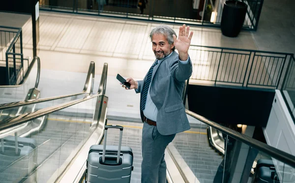 Gülümseyen Olgun İş Adamı 'nın Havalimanında bize el sallamasının portresi. Zarif gri takım elbiseli mutlu bir iş adamı merdivende bavuluyla dururken cep telefonu kullanıyor ve kameraya bakıp veda ediyor..