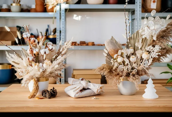 机の上の花器には永遠に2つのドライフラワーの花束がプレゼントされています ワークショップでは 木製のテーブルの上にそれらの間の廃棄物の贈り物と手作りのポットで乾燥した植物と美しい花の配置 — ストック写真