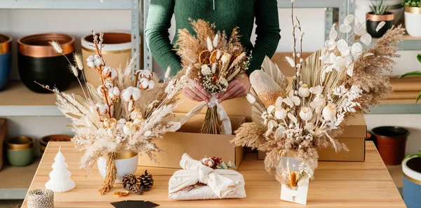 段ボール箱付きのテーブルに立っている間 永遠の乾燥植物や出荷のためのフラワーアレンジメントを保持する女性の手 手作りの花のポットで2花束 プレゼントをラップし ノートありがとう — ストック写真