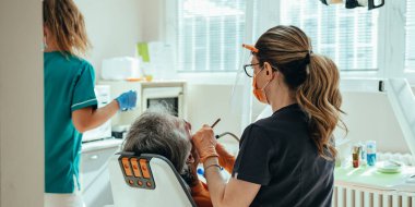 Koruyucu iş giysisindeki tanınmayan kadın diş hekimi, arka planda kadın diş asistanı ve diş sağlığı kliniğindeki erkek dişlerin diş dolgusunu tedavi etmek için diş matkabı kullanıyor.