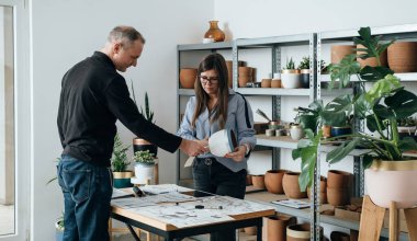 Mobilya Tasarımcısı Ekibi, el yapımı saksılar için bitki tezgahı tasarımında birlikte çalışıyor. Gülümseyen kadın ve erkek meslektaşlar masada durup müşteriler için bir proje tasarımı üzerinde tartışıyorlar. Planlar, dijital tablet ve saksı kullanarak..