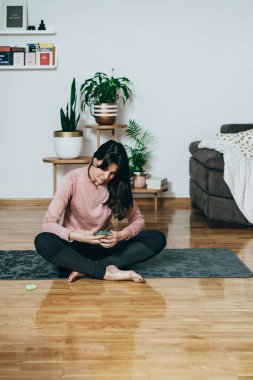 Ev antrenmanından önce cep telefonunu kullanan kablosuz kulaklıklı gülen kadın. Egzersiz minderinde oturan mutlu kadın ve telefonunu internet üzerinden müzik dinlerken bir spor programı seçmek için kullanıyor..