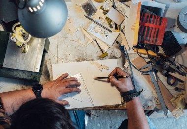 Anonim bir marangoz atölyesinde yeni bir proje üzerinde çalışıyor. Çalışma masasında otururken kalem ve cetvelle yeni parçalar çizen tanınmayan bir adamın masa üstü görüntüsü.